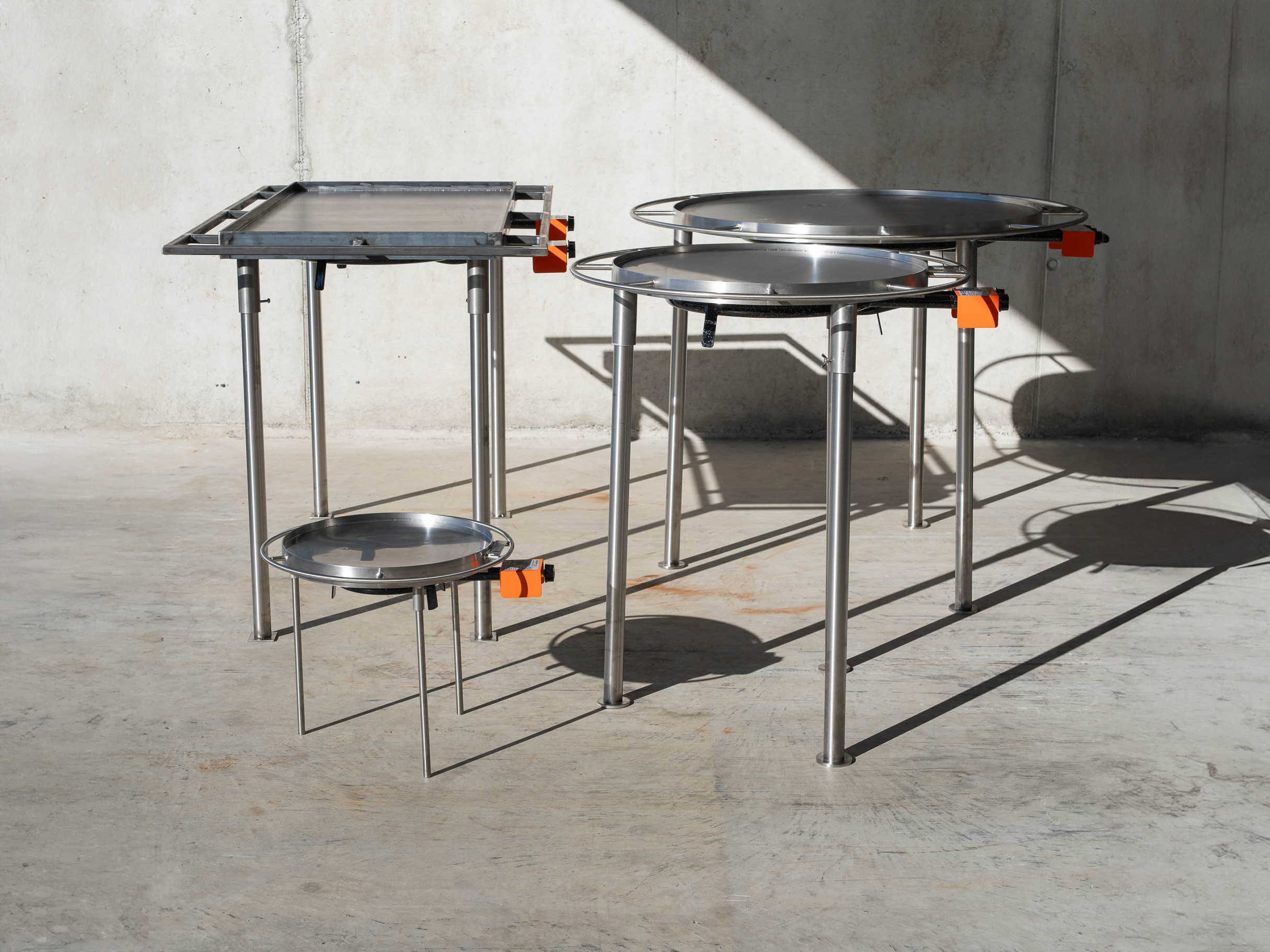 Das Bild zeigt vier verschiedene Gasgrills, sogenannte Mongolengrills, bei denen ein Gasheizkreis eine Metallplatte von unten erhitzt. Drei Grills sind circa Hüfthof, ein kleiner Campinggrill ist ungefähr halb so hoch. Die Grills haben einen Gasregler mit orangener Schutzkappe und rundherum eine Art Reling.